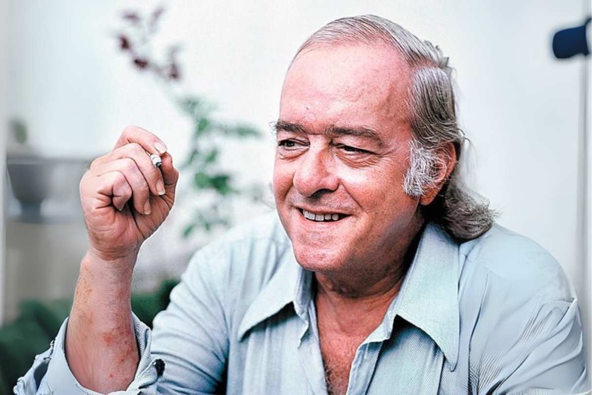 Vinicius de Moraes sorrindo com cigarro na mão, em ambiente iluminado